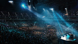 A large concert audience with stage lights and a spotlight on stage in a packed indoor arena.