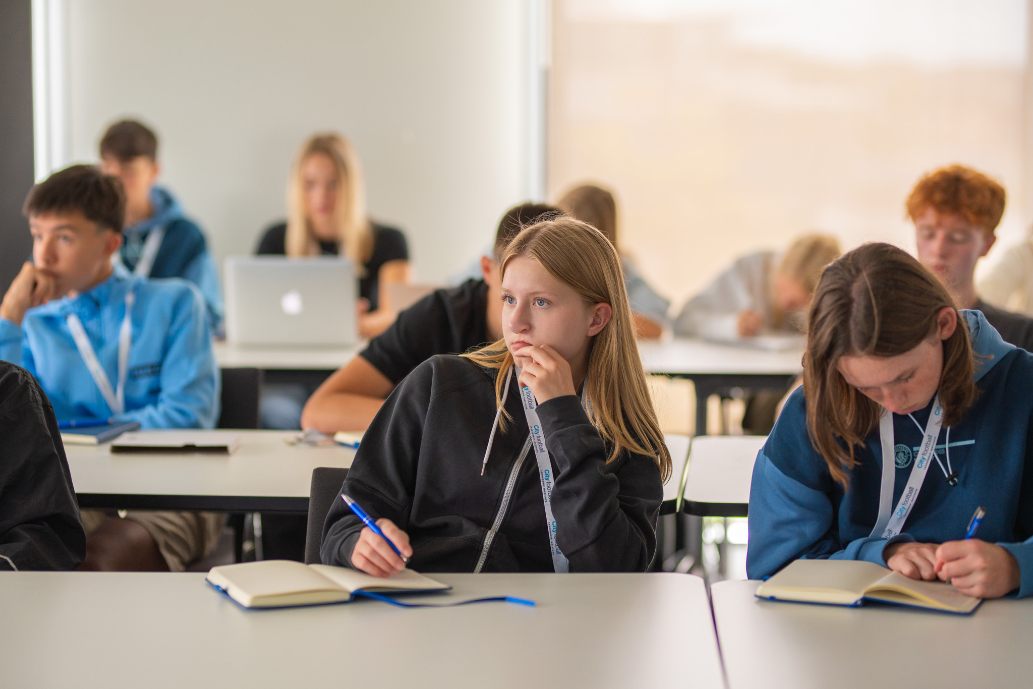A group of students in a classroom setting, sitting at desks with notebooks open. Some are writing while others appear focused on a lecture or discussion.