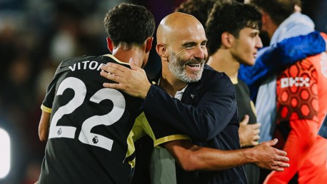 A bald man in a dark jacket hugging a soccer player wearing a black jersey with the number 22 and 'VITOR' on the back. Other people are in the background.
