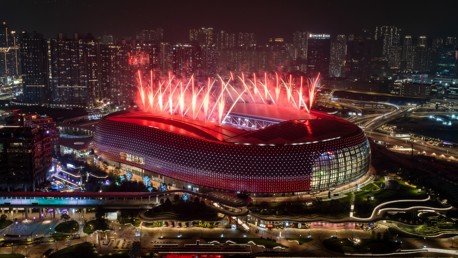 A stadium is illuminated with red lights and surrounded by red fireworks at night in a cityscape setting.