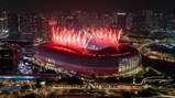 A stadium is illuminated with red lights and surrounded by red fireworks at night in a cityscape setting.