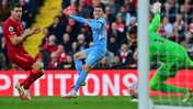 A soccer player in a light blue kit attempts a goal against a team in red. The goalkeeper is wearing a green kit.