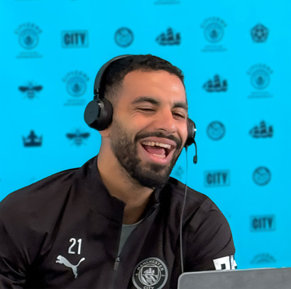 A person in a black Manchester City tracksuit with number 21 and logos, wearing a headset. The background features a blue wall with various Manchester City logos.