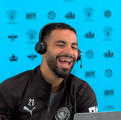 A person in a black Manchester City tracksuit with number 21 and logos, wearing a headset. The background features a blue wall with various Manchester City logos.