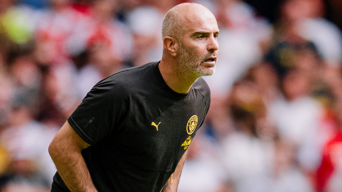 A man wearing a black Man City shirt on the sideline during a football match, focused and gesturing energetically.