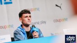 A person holding a microphone at a press conference for the Asia Tour '26. The background displays logos and text including 'Coupang' and 'Puma'.