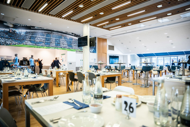 The image shows a spacious hospitality area at Etihad Stadium with tables set for dining. A large stadium mural decorates the wall, staff members are visible, and seating arrangements include chairs and bar stools.