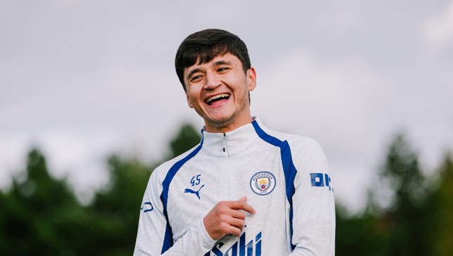 A person wearing a Manchester City training jacket, smiling in an outdoor setting with trees in the background.
