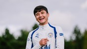 A person wearing a Manchester City training jacket, smiling in an outdoor setting with trees in the background.