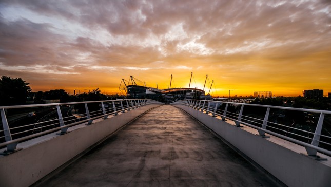 A view of the Etihad Stadium at sunset, taken from a pedestrian bridge leading to the stadium.