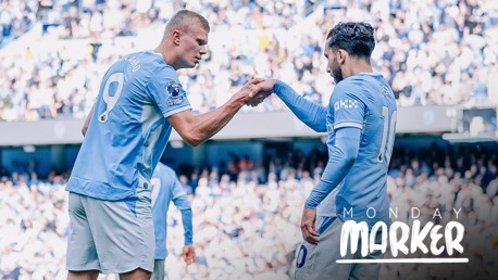 Two Manchester City players in blue jerseys are on the field performing a fist bump. The text 'Monday Marker' is visible in the bottom right corner.