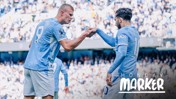 Two Manchester City players in blue jerseys are on the field performing a fist bump. The text 'Monday Marker' is visible in the bottom right corner.