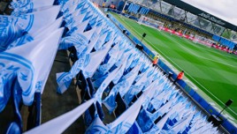 Empty stadium seats lined with blue and white Manchester City flags overlooking the Joie Stadium field.