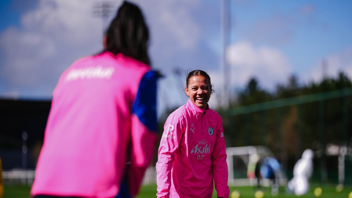Two players wearing pink Manchester City training kits practice on a football field on a clear day.