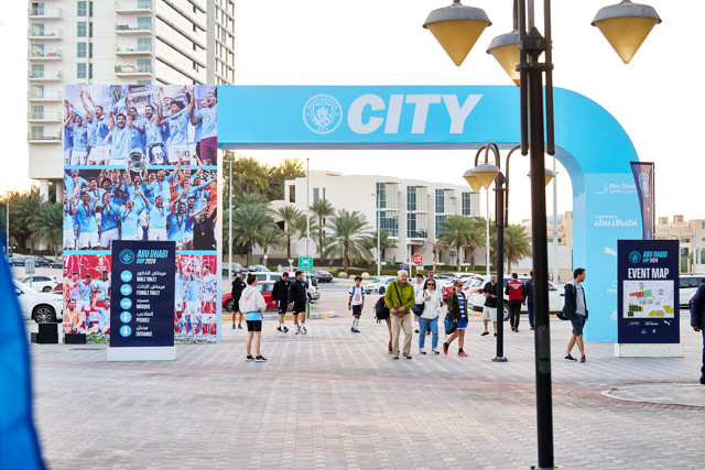 Entrance gate with a 'CITY' banner and event signs in Abu Dhabi. Manchester City branding, event map stand, and people walking around the area. Posters of football celebrations displayed on the side.