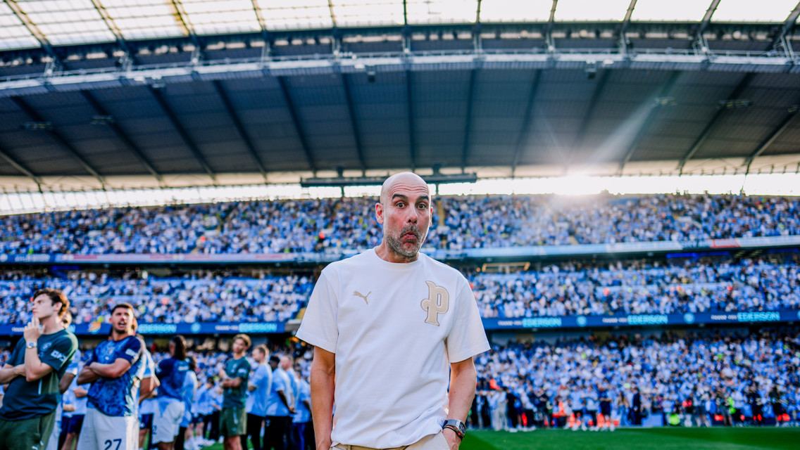 A man wearing a white Puma shirt walks on a green field with a crowded football stadium in the background. Sunlight shines from above.