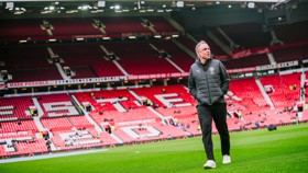 A person walking across a football field in a stadium with red seats, possibly Manchester United's stadium.