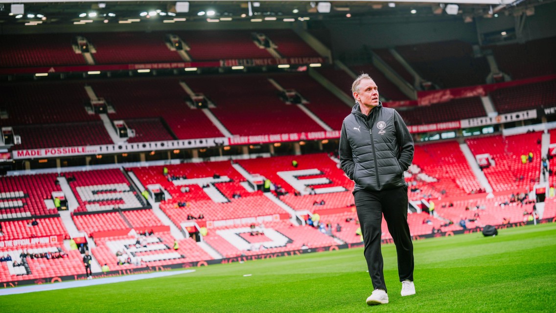 A person walking across a football field in a stadium with red seats, possibly Manchester United's stadium.