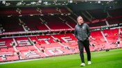 A person walking across a football field in a stadium with red seats, possibly Manchester United's stadium.
