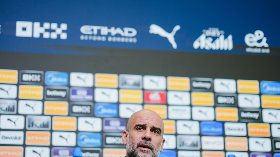 A blurred person stands in front of a press conference backdrop featuring multiple brand logos including Etihad, Puma, and OKX.