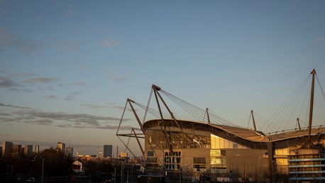 View of the Etihad Stadium in Manchester at sunset with city skyline in the background.