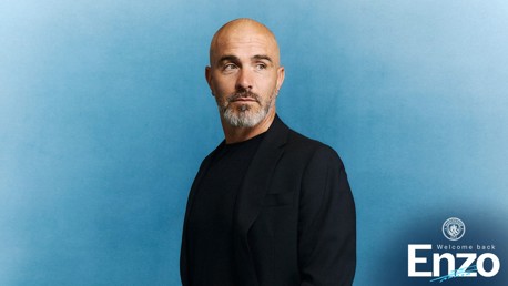 A man with a bald head and grey beard wearing a black jacket against a blue background. Text reads 'Welcome back Enzo' with a Manchester City logo.