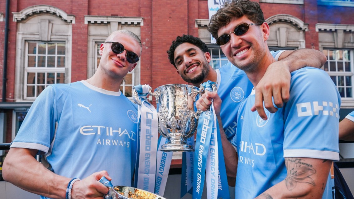 Three individuals wearing Manchester City jerseys hold a Carabao Cup trophy with ribbons in a city setting.