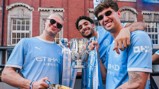 Three individuals wearing Manchester City jerseys hold a Carabao Cup trophy with ribbons in a city setting.