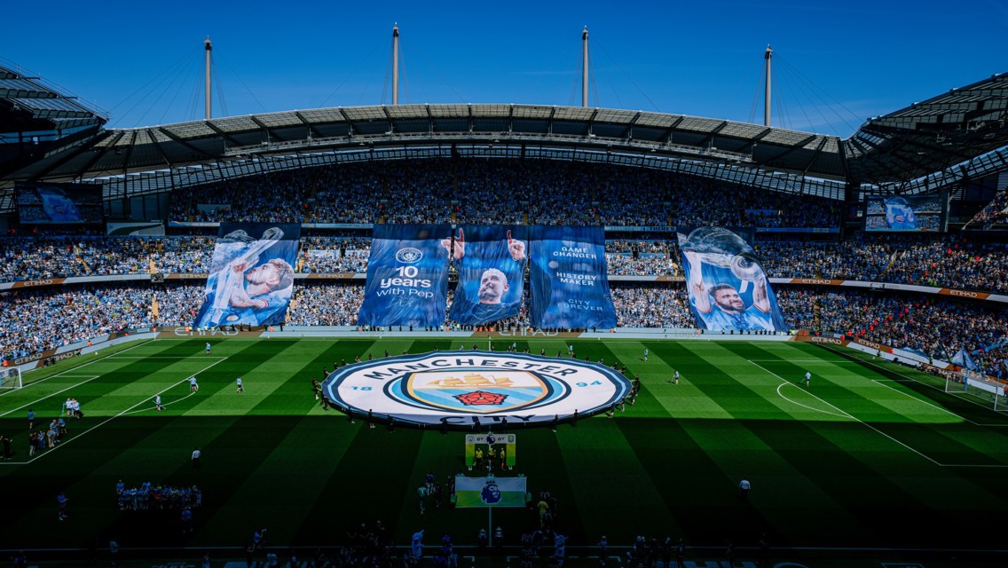 A wide view of Manchester City's Etihad Stadium with large banners celebrating '10 Years with Pep'. The stadium is filled with fans and features the Manchester City logo on the pitch.