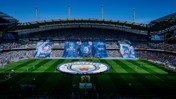 A wide view of Manchester City's Etihad Stadium with large banners celebrating '10 Years with Pep'. The stadium is filled with fans and features the Manchester City logo on the pitch.
