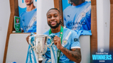 A person in a Manchester City jersey holding the Carabao Cup trophy with 'Winners 2026' text in the corner.
