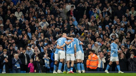 Manchester City players celebrate a goal in front of cheering fans in the Etihad Stadium. One player walks away while others embrace in a group hug. Many spectators are capturing the moment on their phones.
