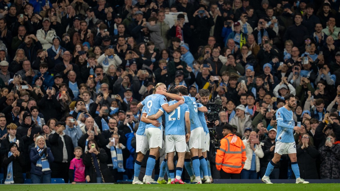 Manchester City players celebrate a goal in front of cheering fans in the Etihad Stadium. One player walks away while others embrace in a group hug. Many spectators are capturing the moment on their phones.