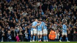 Manchester City players celebrate a goal in front of cheering fans in the Etihad Stadium. One player walks away while others embrace in a group hug. Many spectators are capturing the moment on their phones.