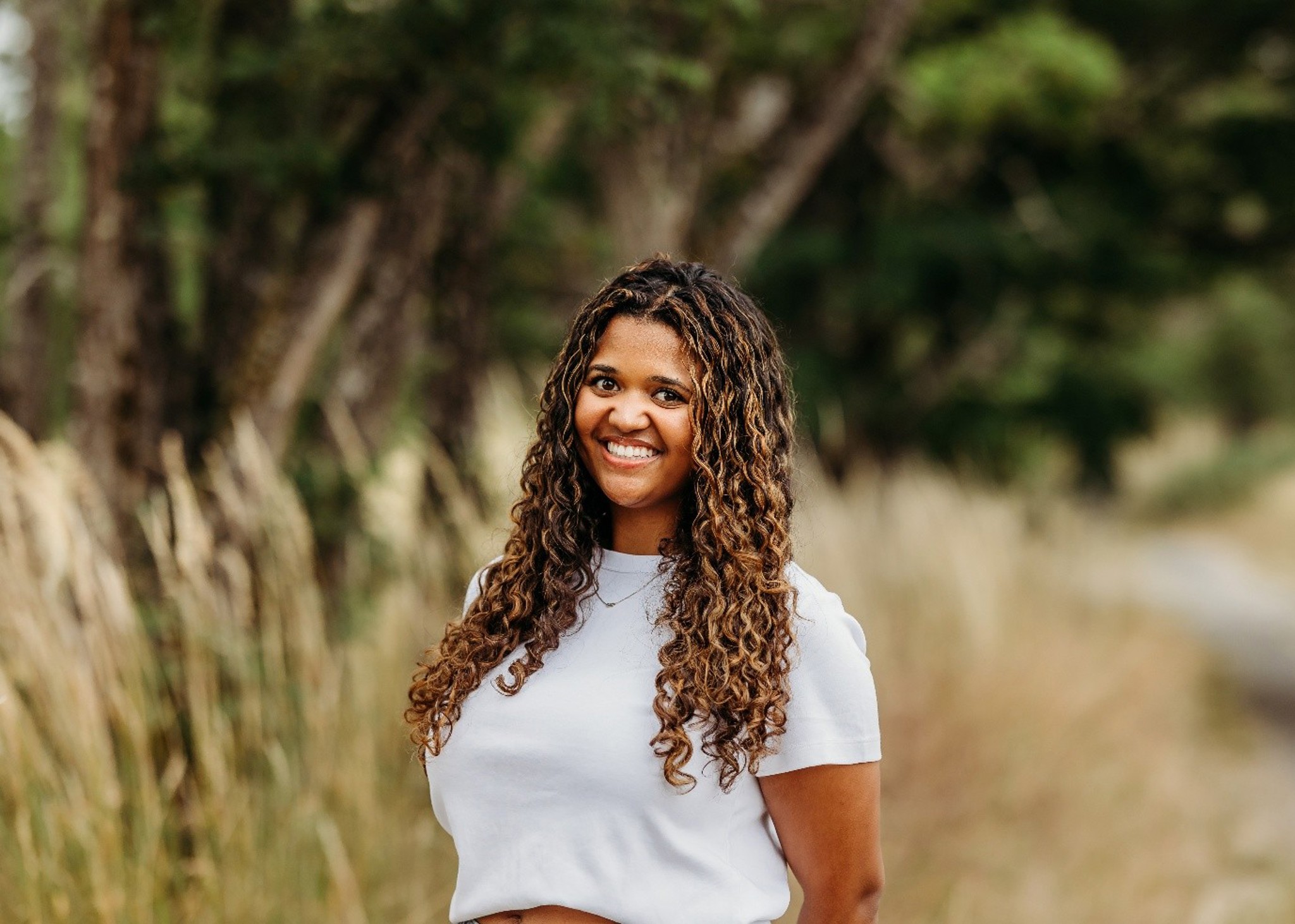 A woman with curly hair smiles while standing outdoors. She is wearing a white t-shirt and surrounded by tall grass and trees.