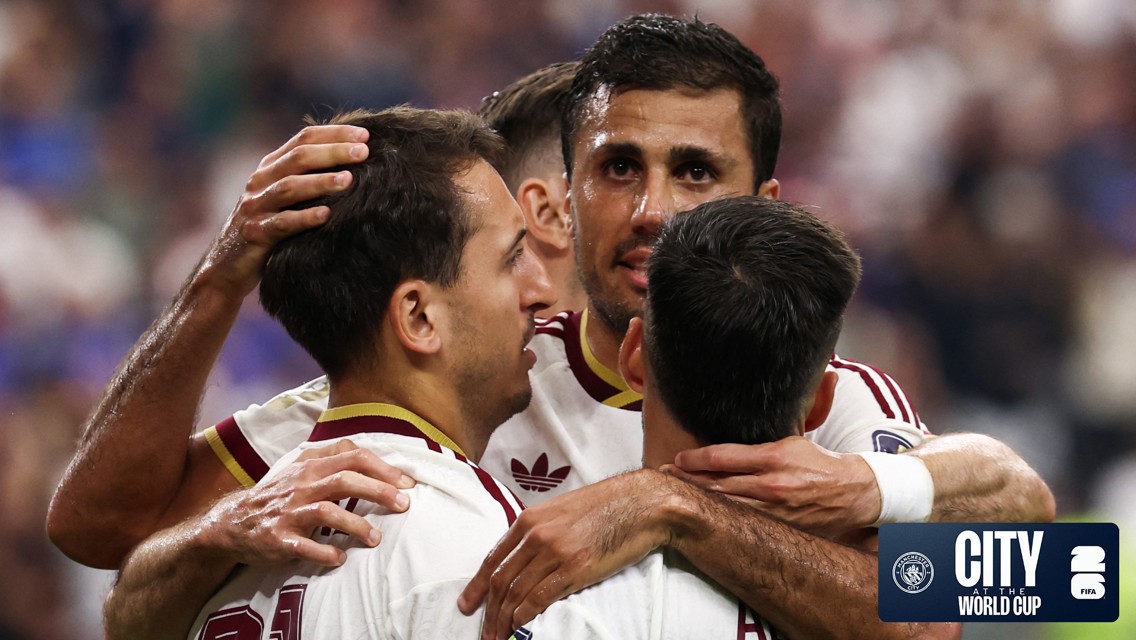 Football players in white jerseys embracing on the field in celebration. Manchester City logo and World Cup text in the corner.