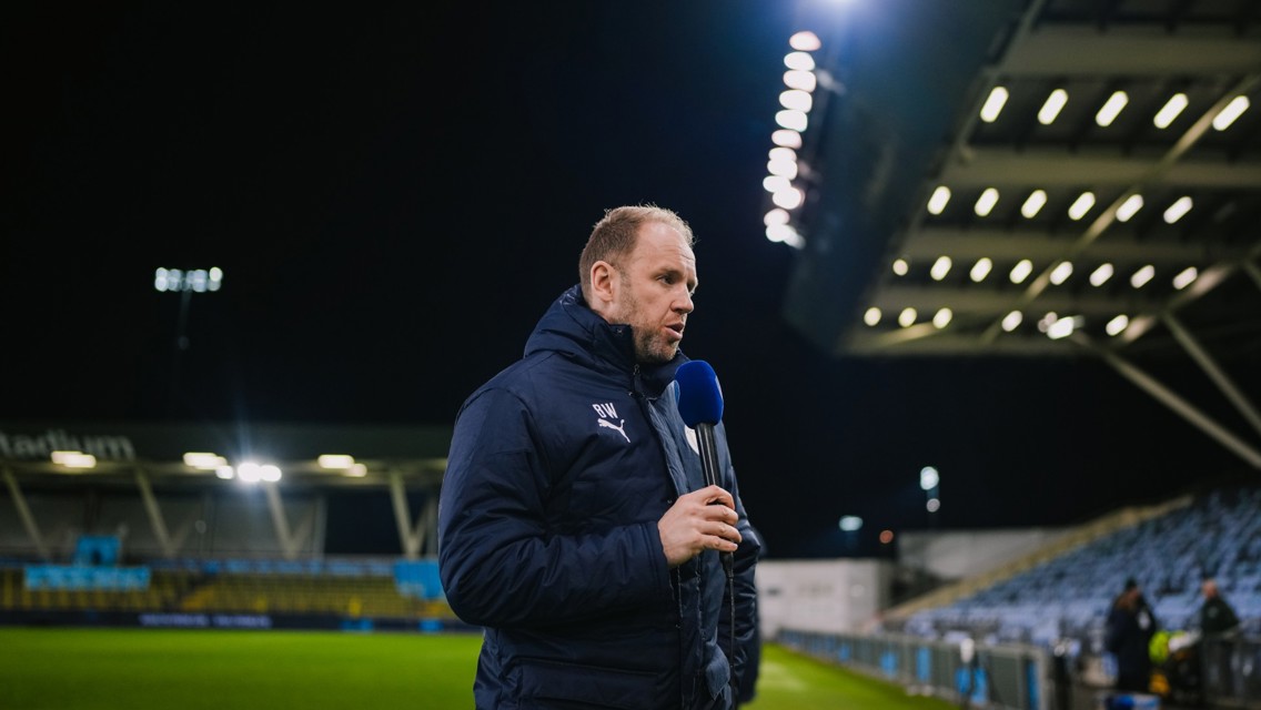 A person holding a microphone stands on the side of a football field in a stadium at night, possibly conducting an interview or broadcast.