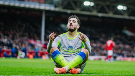 A Manchester City player sitting on the grass, wearing a gray vest and neon socks during a match. The player's face is blurred, and the stadium is in the background.