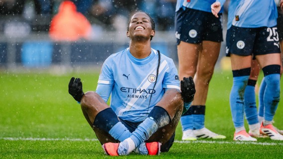 Manchester City player in light blue kit sitting in a meditation pose on the field during a match, with blurred face