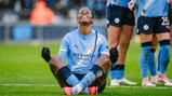 Manchester City player in light blue kit sitting in a meditation pose on the field during a match, with blurred face