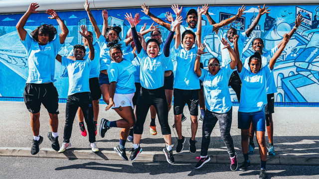A group of people jumping joyfully in blue t-shirts with 'CITY' visible, in front of a blue mural wall, suggesting a team event or celebration.