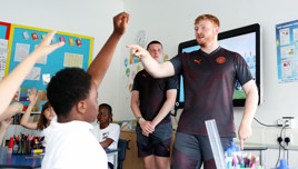 Two individuals wearing Manchester City apparel lead an interactive session with children in a classroom setting. One individual is pointing while children with raised hands are engaged.