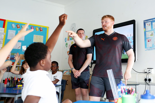 Two individuals wearing Manchester City apparel lead an interactive session with children in a classroom setting. One individual is pointing while children with raised hands are engaged.