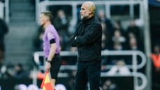 A football coach is standing on the sideline during a match, wearing a black jacket and black pants. A referee in a purple shirt is walking nearby.