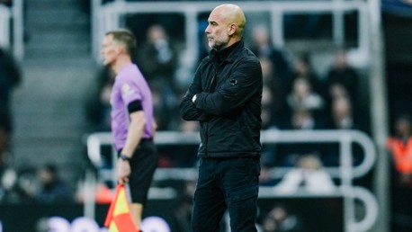 A football coach is standing on the sideline during a match, wearing a black jacket and black pants. A referee in a purple shirt is walking nearby.