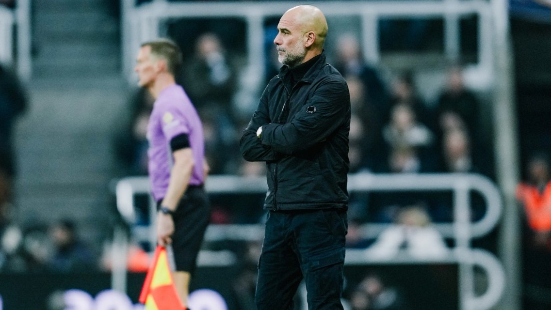 A football coach is standing on the sideline during a match, wearing a black jacket and black pants. A referee in a purple shirt is walking nearby.