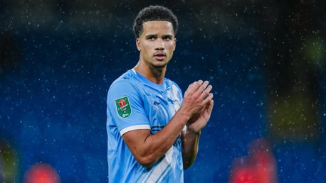 A football player in a light blue kit is clapping in the rain. The kit has a Carabao Cup patch on the sleeve.