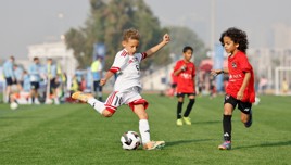 Two young players in soccer jerseys, one in white and the other in red, engaged in a soccer match on a green field. The player in white is attempting a kick.