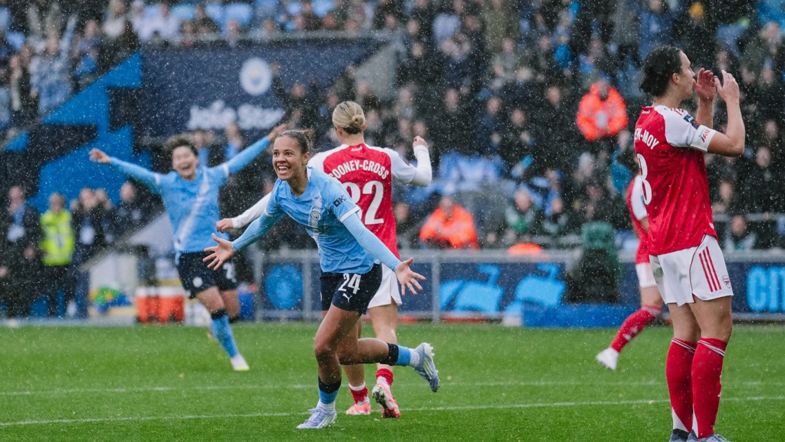 Football players in blue and red jerseys celebrate a goal in rainy weather on a crowded stadium field.