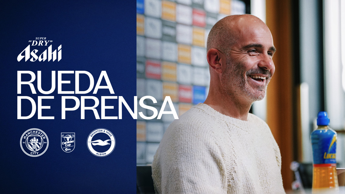 A person smiles during a press conference with Manchester City and Brighton & Hove Albion logos. 'Rueda de Prensa' text is visible.